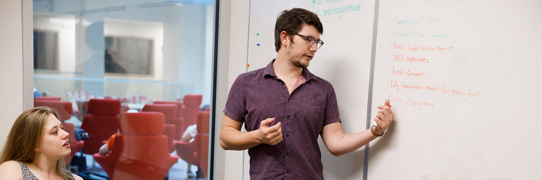 A student writes on a whiteboard in a group meeting room as one of his group members watches.