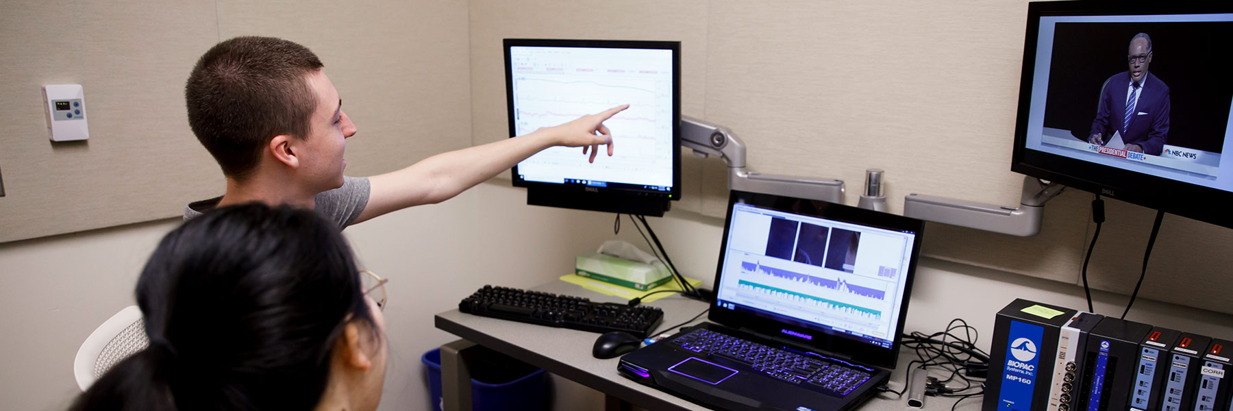 Two students record data for research while they watch a reporter on a screen.