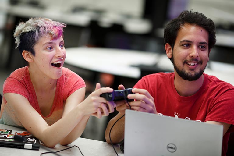 Two students interact with a game controller and a laptop at a student club event.