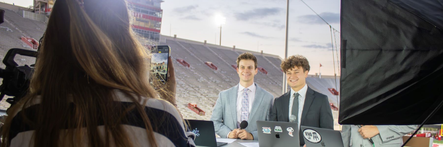 Two reporters holding microphones stand smiling as a person takes a picture of them with their phone with several cameras off to the side and an empty IU football stadium behind the students.