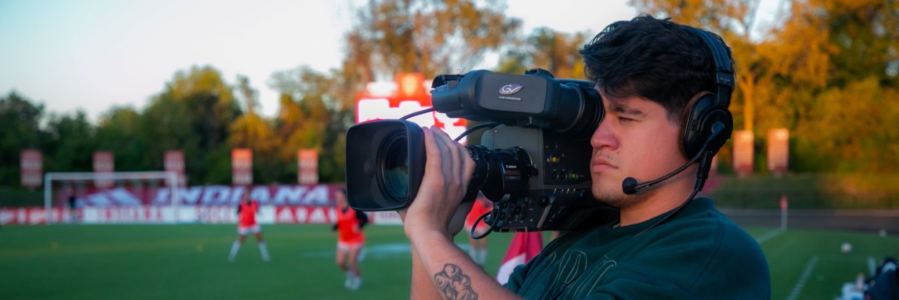 Person wearing a headset and holding a video camera on their shoulder while standing on a soccer field with several players in the background.