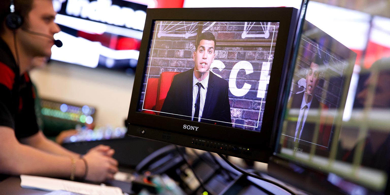Students working in a TV production studio watch a video of a reporter speaking on a monitor.
