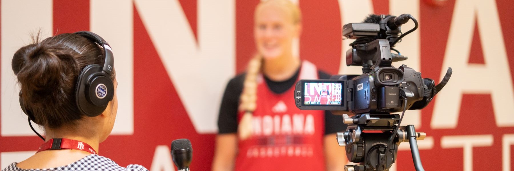 Person holding a microphone with a video camera recording on a tripod in the forefront with a student athlete standing in the background along a wall that spells "INDIANA."