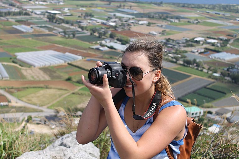 A woman takes a photo standing on a hillside above a valley.