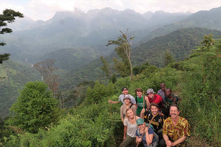 A group of students stands on an overgrown hillside over a valley.